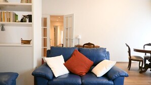 Een blauwe bank met kussens, een boekenkast, een eettafel en een stoel in een goed verlichte kamer.