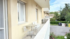 Een balkon met een tafel en stoelen, een afvalbak, en een parkeerplaats met een auto en groen.