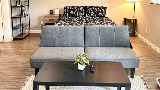 A living room with a grey sofa, a coffee table with a plant, a bookshelf, and a bed with floral bedding.