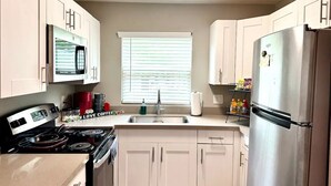 A kitchen with white cabinets, a stainless steel refrigerator, a microwave, a stove, and a window with blinds.