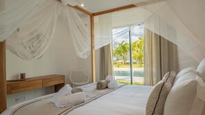Un dormitorio con una cama grande, una mesita de noche de madera, una silla y vista a una piscina y palmeras a través de una puerta de vidrio.