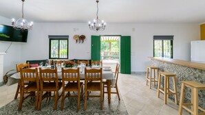 Comedor con una mesa dispuesta para cuatro, sillas de madera, una araña y un televisor montado en la pared, con vistas al exterior a través de una puerta.