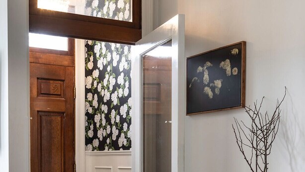 Eine Holztür mit Glasfenster, eine weiße Tür mit Glasfenster, floraler Schwarz-Weiß-Tapete, ein gerahmtes Bild und eine Vase mit Zweigen auf einem Tisch.