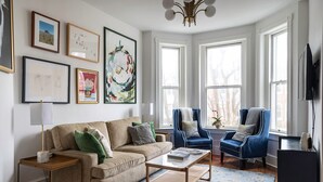 Ein Wohnzimmer mit einem beigefarbenen Sofa, zwei blauen Sesseln, einem Couchtisch und einer TV-Konsole. An der Wand befinden sich gerahmte Bilder und ein Kronleuchter.