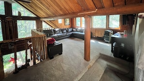 A cozy cabin interior with a wooden ceiling, a leather sofa, a fireplace, and large windows offering a view of the forest.