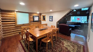 A cozy log cabin interior with a wooden dining table, chairs, and a large window overlooking a snowy landscape.