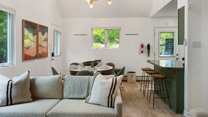 A modern living room with a sofa, a dining area with a table and chairs, a kitchen area with stools, and a chandelier.