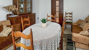 Un comedor con una mesa redonda cubierta por un mantel a cuadros, rodeada de sillas de madera. Se ve un aparador con puertas de cristal a la izquierda, y una puerta al fondo.