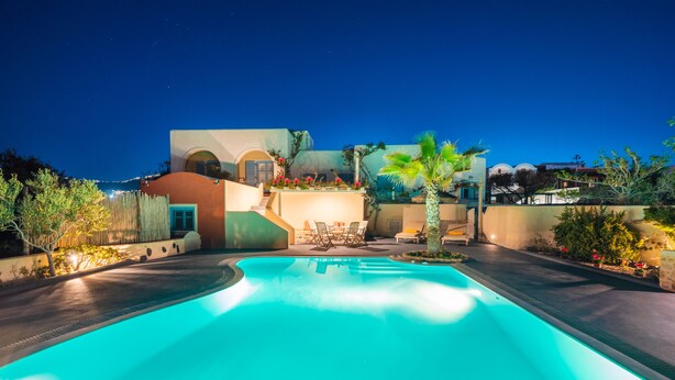 A swimming pool with a curved edge, surrounded by a landscaped area with palm trees and a building with a curved roof and arched windows. - Daphne | Cycladic Three-Bedroom Villa with Private Pool and Sea View (Thira - Santorini)