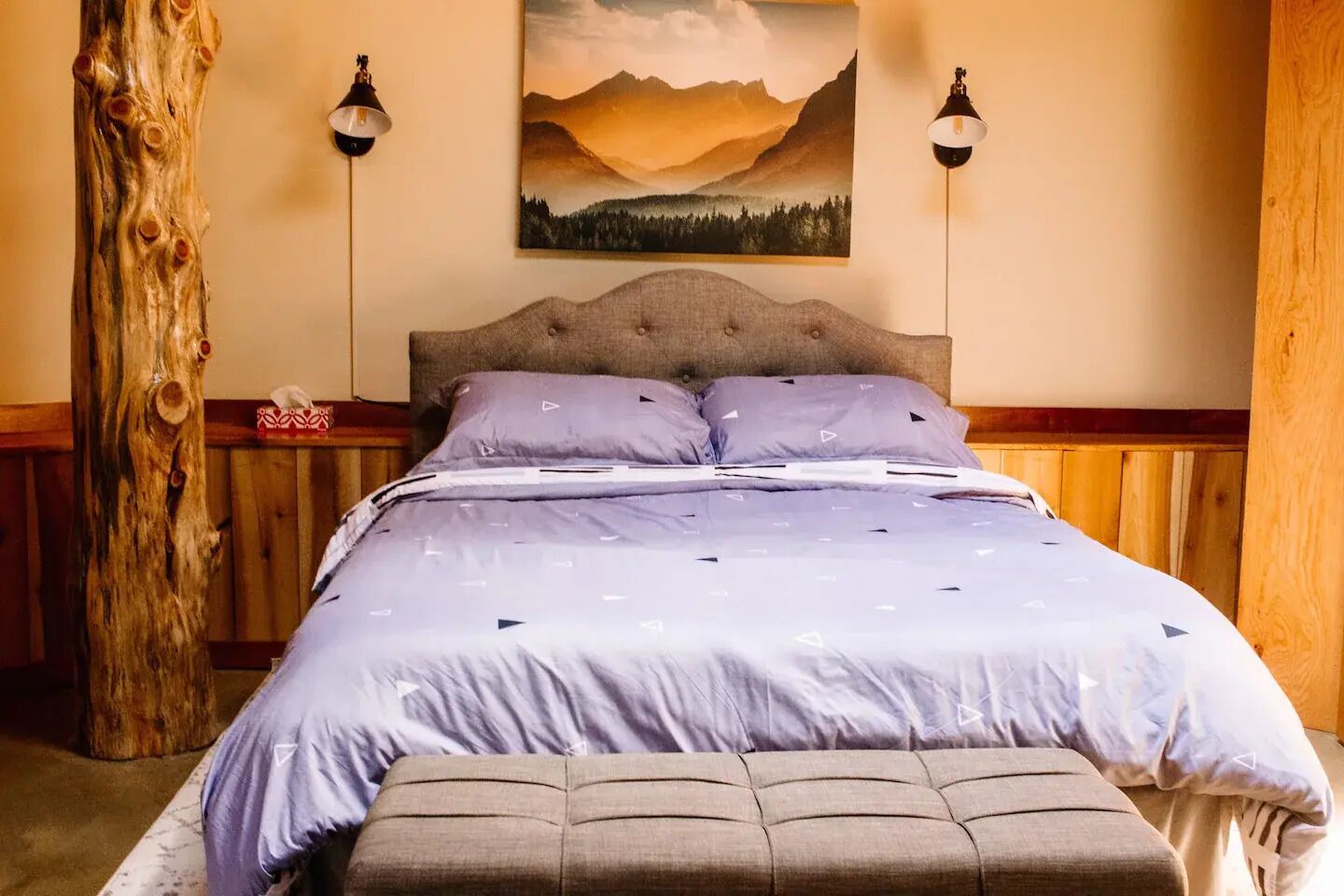 Un dormitorio con una cama, dos lámparas de pared, un cuadro de montañas y un tronco de árbol de madera.