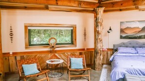 Une pièce confortable avec un lit, deux fauteuils en osier, une petite table, une fenêtre donnant sur la verdure et un tableau accroché au mur.
