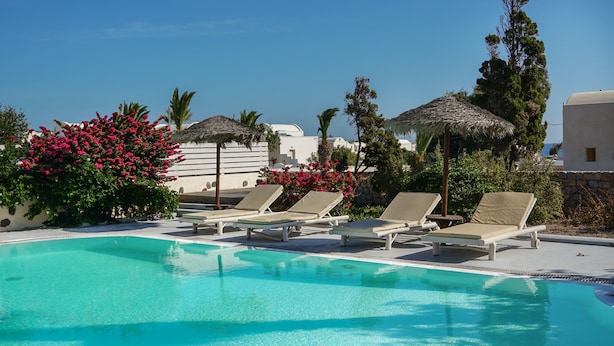 Piscina azul límpida com espreguiçadeiras e toldos de palha, rodeada por vegetação e edifícios brancos.