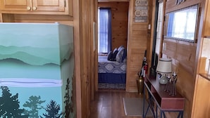 Ein Ferienhausinnenraum mit Holzboden, einem Bett mit gemusterten Betttextilien, einem Kühlschrank mit Landschaftsdarstellung, einem Tisch mit Lampe und einem Schild mit der Aufschrift 'CABIN'.
