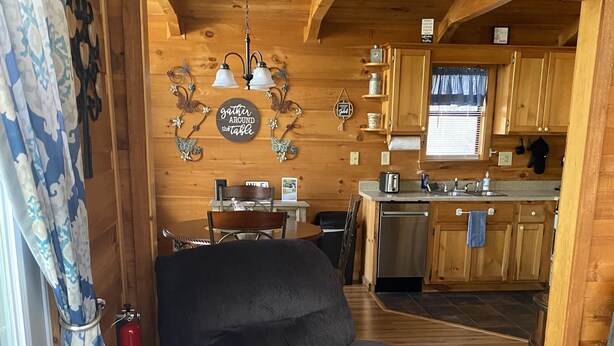 Ein gemütliches Blockhaus-Interieur mit einer Essgegend aus Holz, einer Küche mit Holzschränken und einem bequemen grauen Sessel an einem Fenster mit gemusterten Vorhängen.