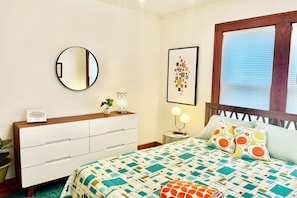 A bedroom with a bed, a dresser, a round mirror, a framed picture, and a window with blinds.