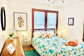Ein ordentlich eingerichtetes Schlafzimmer mit einem Bett, Nachttischlampen, einem Nachttisch mit Pflanze und einem gerahmten Kunstwerk an der Wand.