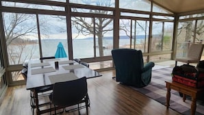 A room with a large window overlooking a body of water, a dining table with chairs, a green armchair, and a small table with a bag. - Waterfront Stay at Sandpiper Cove (Eufaula)