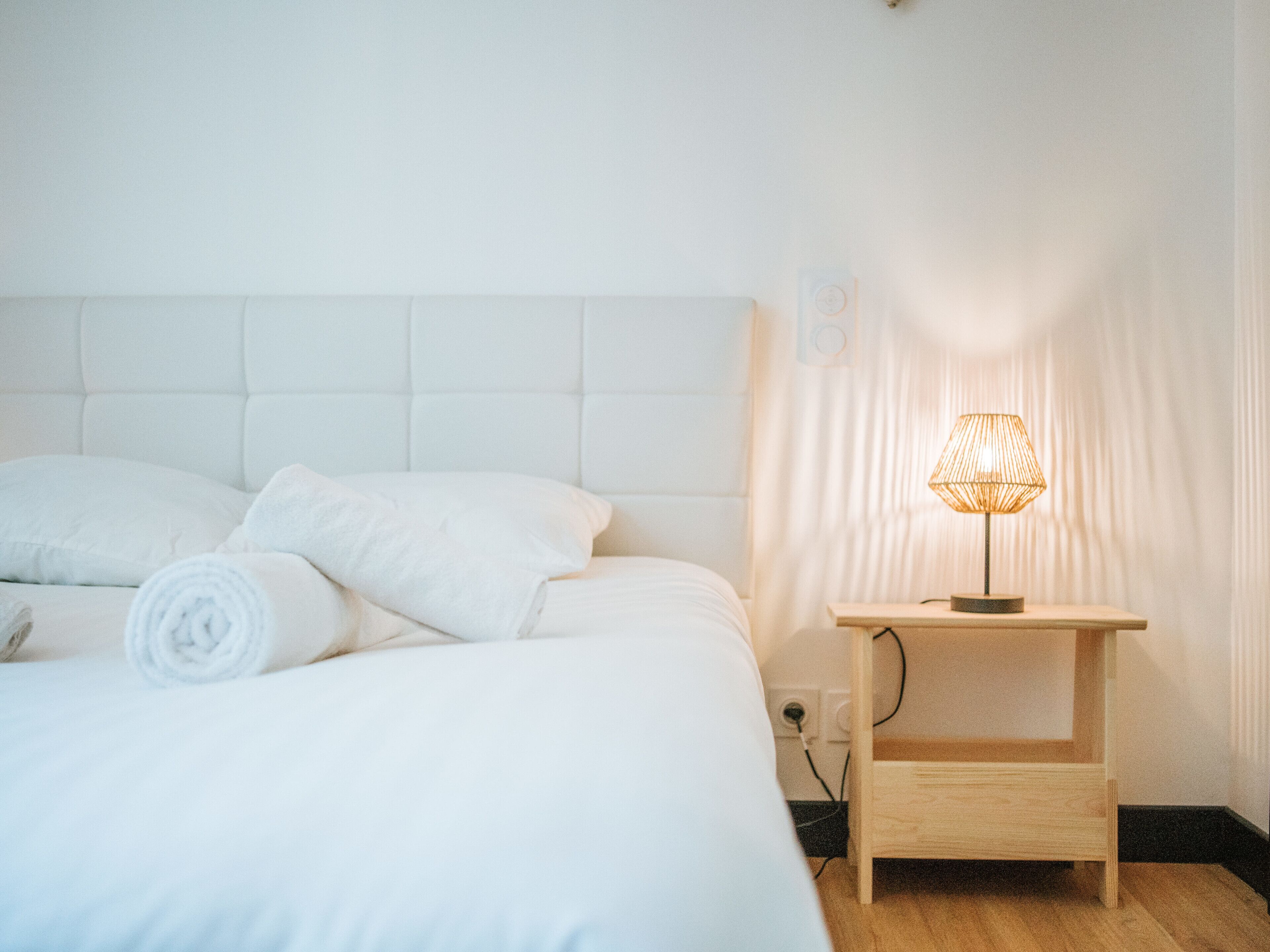 Cama bem arrumada com roupa branca e toalha enrolada, mesa de cabeceira com abajur e tomada na parede.