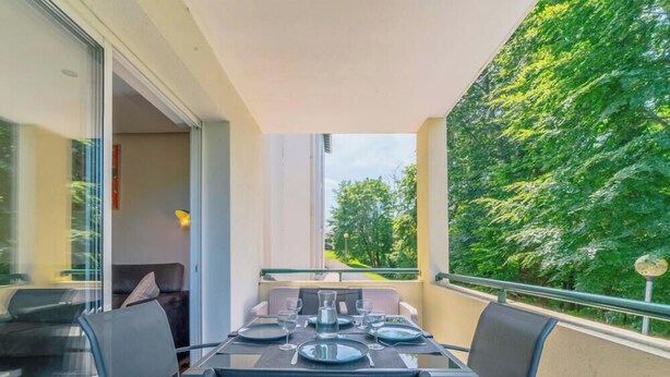 A balcony with a dining table set for two, chairs, and a view of a green tree-lined area. - Le Tarnosien - Landes - Côte Basque (Tarnos)