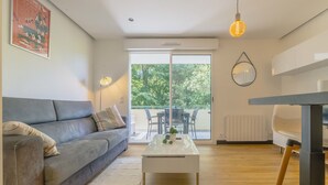 A modern living room with a grey sofa, a white coffee table, and a dining area with a table and chairs. There is a large window with a view of trees outside. - Le Tarnosien - Landes - Côte Basque (Tarnos)