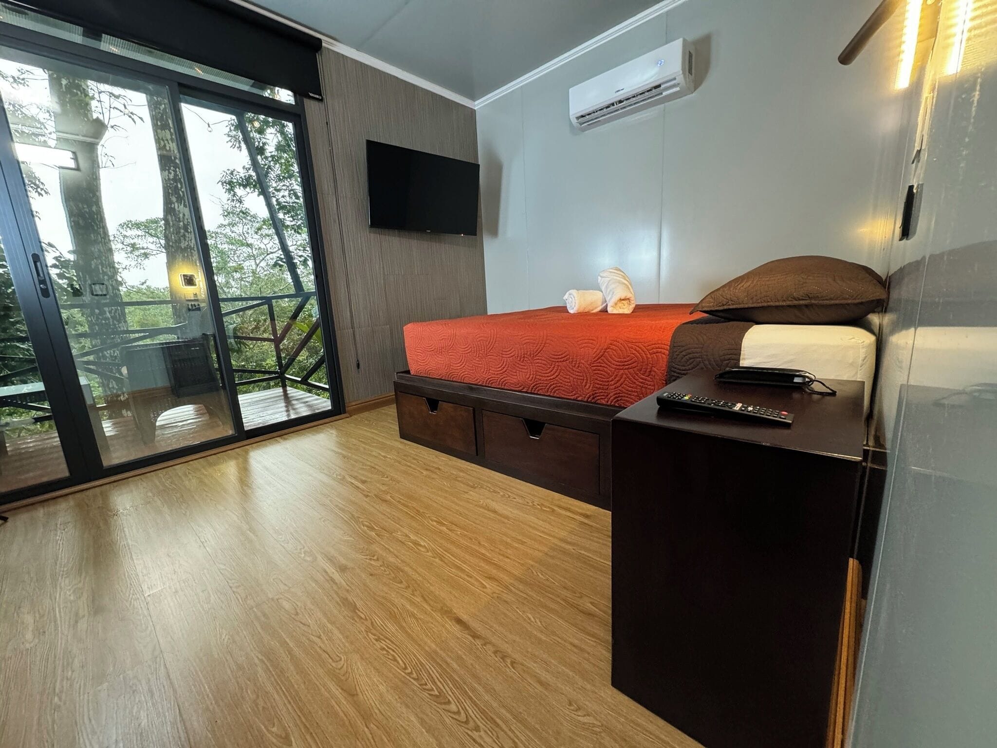 A bedroom with a bed, a TV, an air conditioner, and a sliding glass door to a balcony.