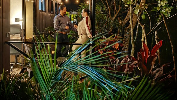 A modern outdoor seating area with wooden beams and string lights, surrounded by lush greenery and plants.