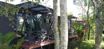 A modern outdoor seating area with a glass enclosure, surrounded by lush greenery and trees.