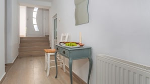 Un couloir avec un escalier, une chaise blanche, une table bleue avec une assiette de raisins et un radiateur blanc.