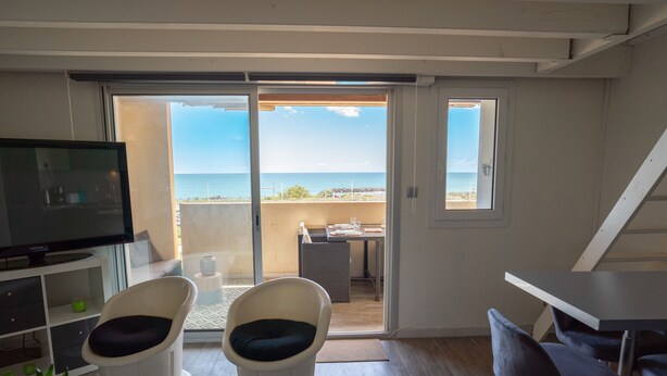 A room with a TV, two chairs, a table, and a view of the ocean through a sliding door.