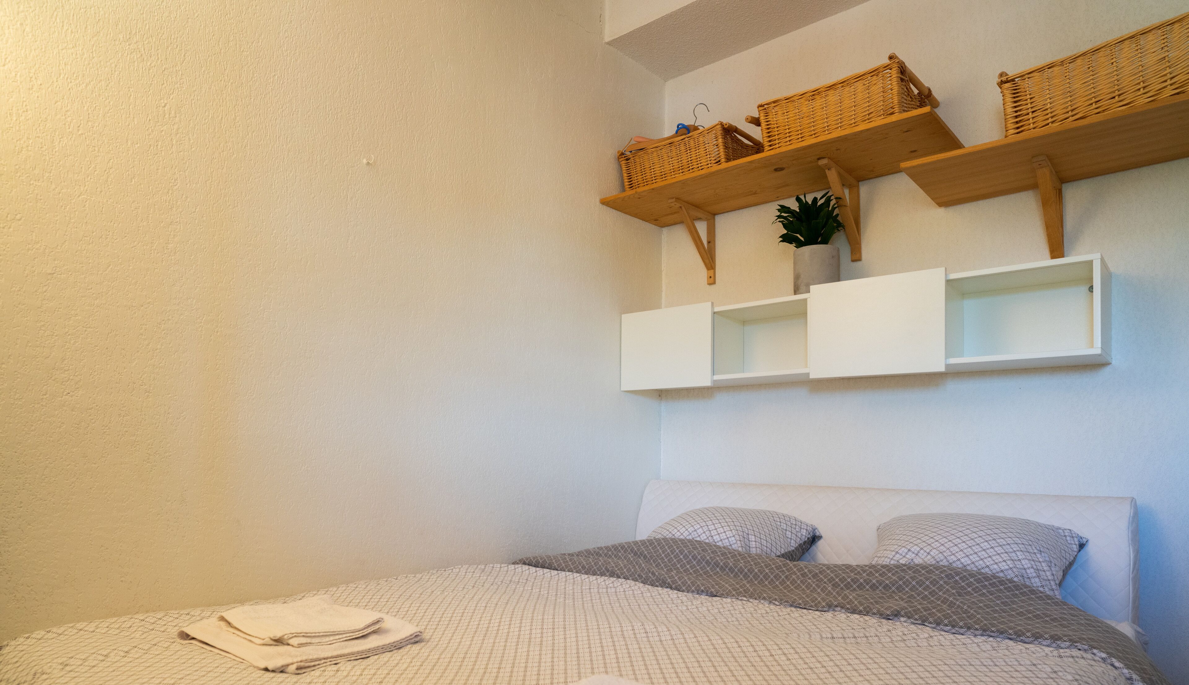 Un dormitorio con una cama, dos almohadas, una toalla doblada y dos cestos de mimbre en un estante de madera sobre la cama.