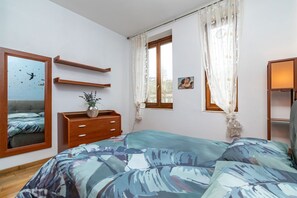 Un dormitorio con cama, cómoda, espejo, ventana con cortinas y estante fijado a la pared.