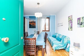 Una habitación con una puerta verde azulada, una mesa de comedor con sillas, un sofá, un lámpara colgante y una pared con una pintura de un paisaje urbano.