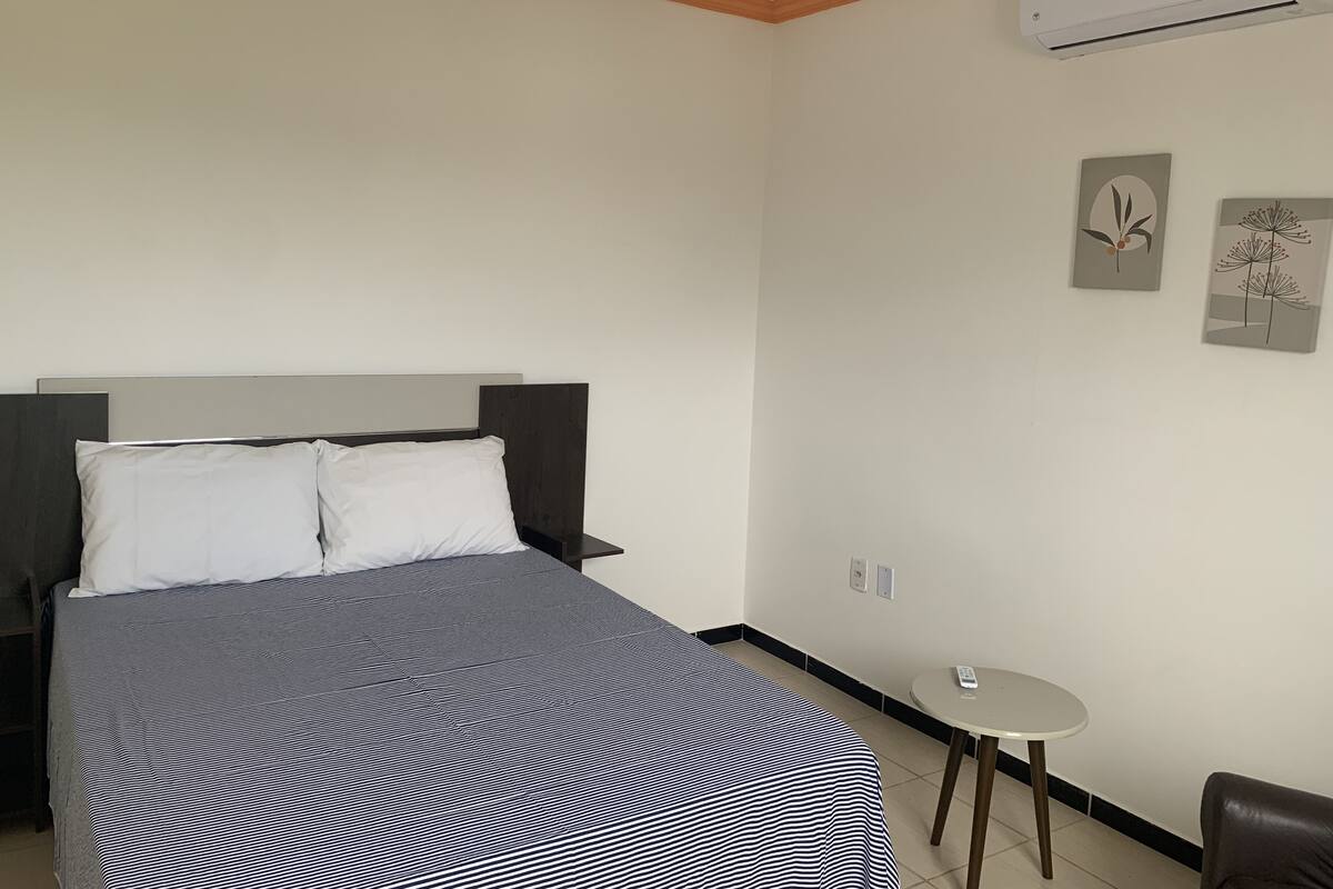 A hotel room with a bed, a small table, a chair, and an air conditioning unit on the wall.