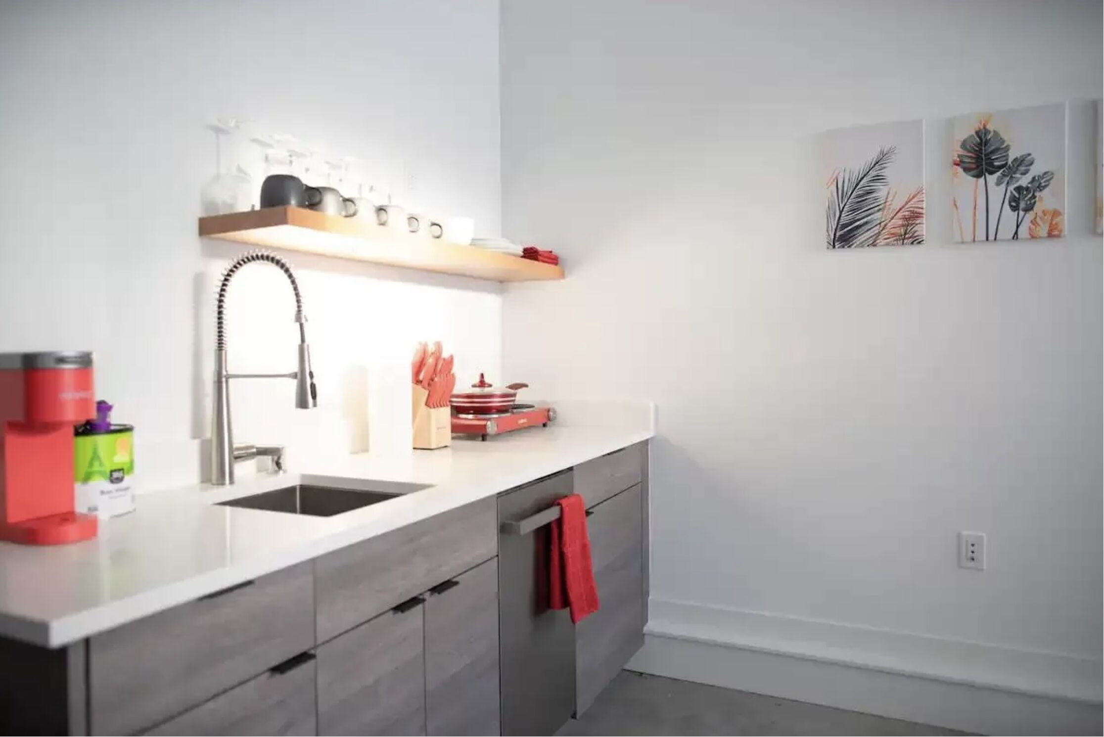 Une cuisine moderne avec un plan de travail blanc, un évier en acier inoxydable et un four encastré. On y trouve des ustensiles de cuisine et un torchon rouge accroché à l’armoire.