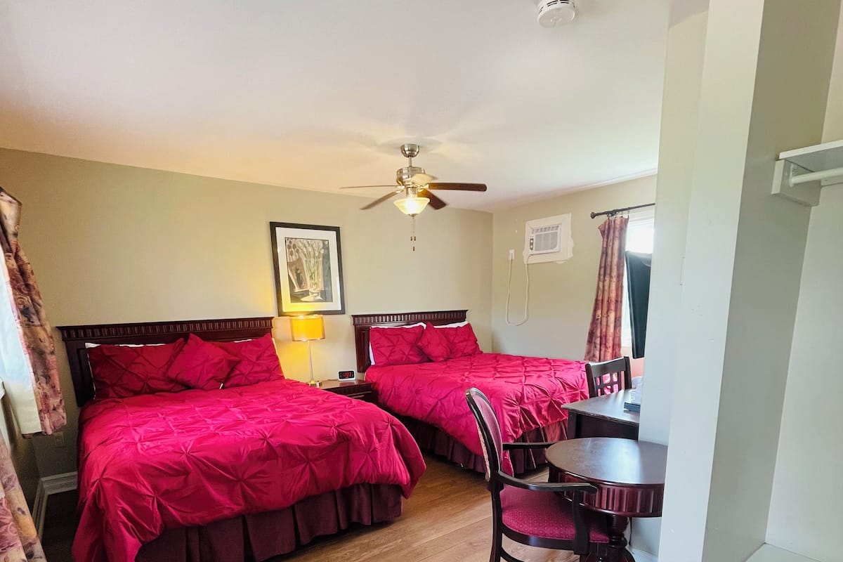 A room with two beds, a ceiling fan, a table with chairs, and a wall-mounted air conditioning unit.