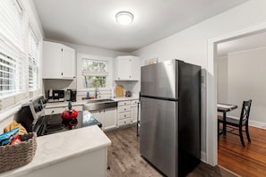 A compact kitchen with white cabinets, a stainless steel refrigerator, a red kettle, and a dining area with a wooden table and chair.
