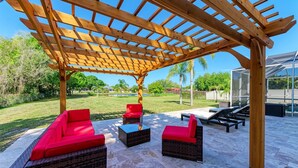 A covered outdoor seating area with wicker furniture and red cushions, a pergola overhead, and a view of a green lawn and a house in the background.