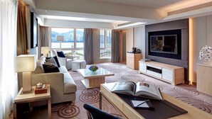 Een moderne hotelkamer met een bank, fauteuil, salontafel, televisie en uitzicht op de stad door grote ramen.