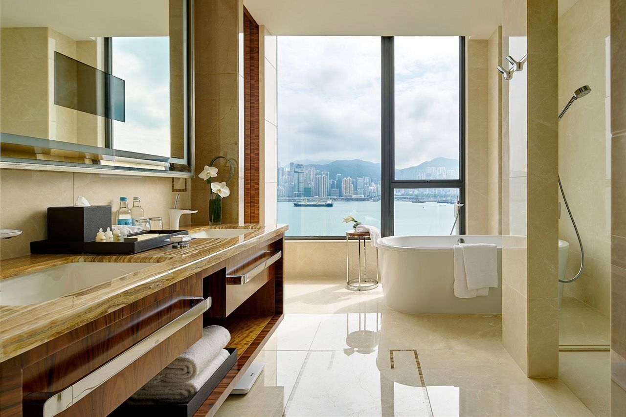 Un baño moderno con un espejo grande, un tocador de madera, una bañera y vistas a la ciudad a través de amplias ventanas.