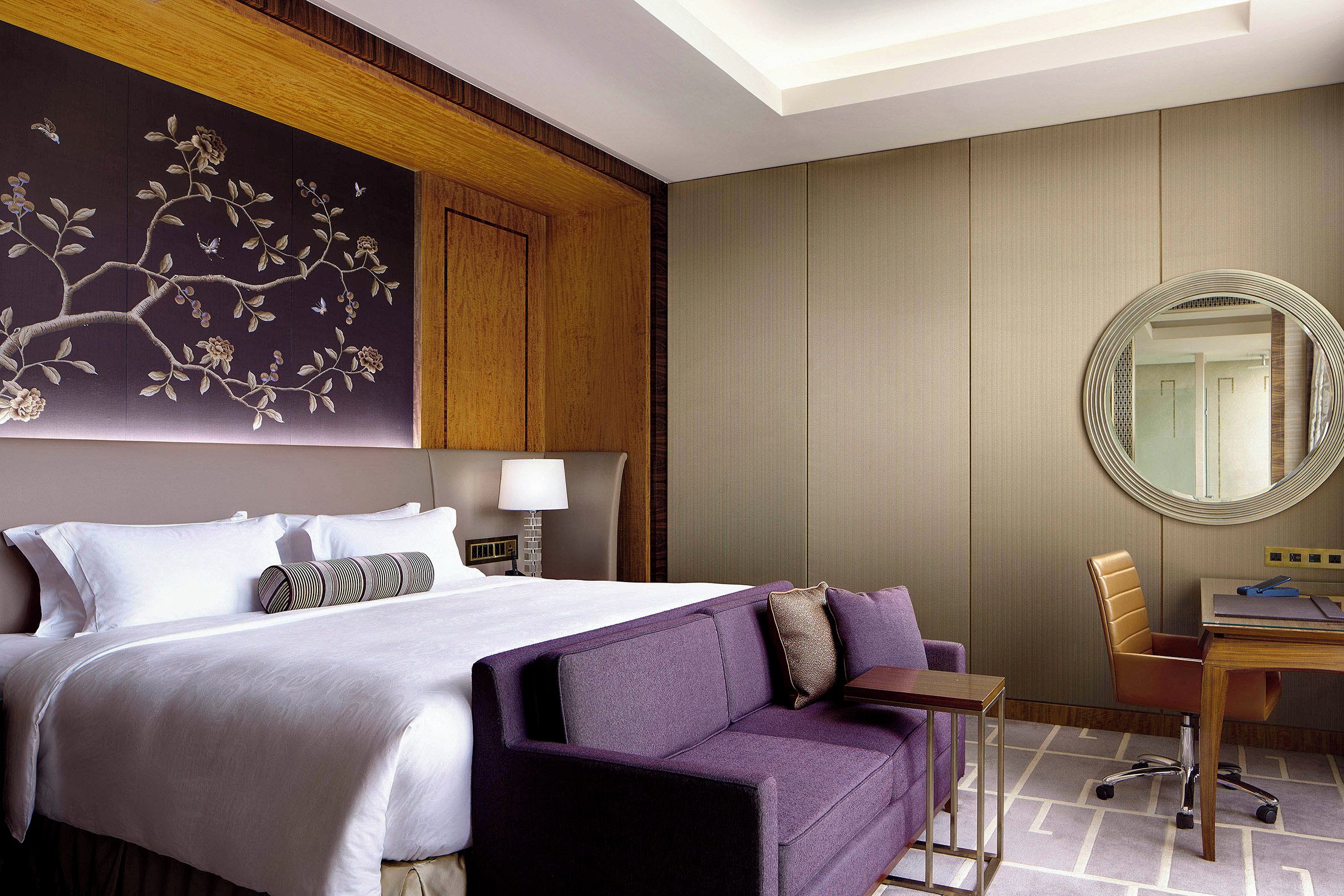 A hotel room with a large bed, a purple sofa, a desk, and a mirror.