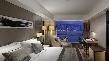 A hotel room with a large bed, a desk, a chair, a view of the cityscape, and a window with curtains.