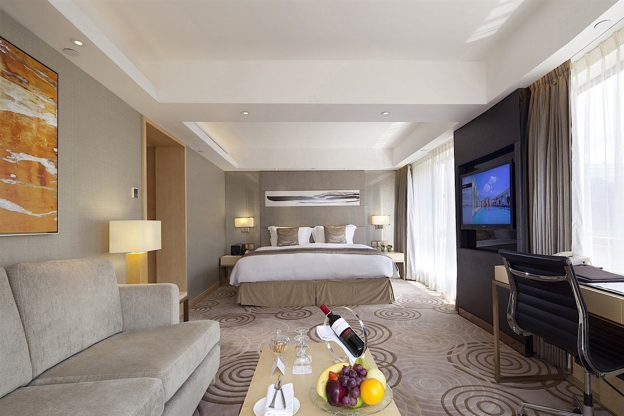 A hotel room with a bed, a TV, a desk, a chair, a sofa, and a table with fruit and wine.