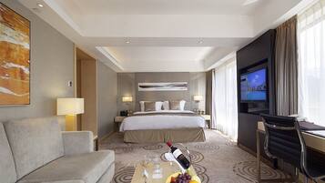 A hotel room with a bed, a TV, a desk, a chair, a sofa, and a table with fruit and wine.