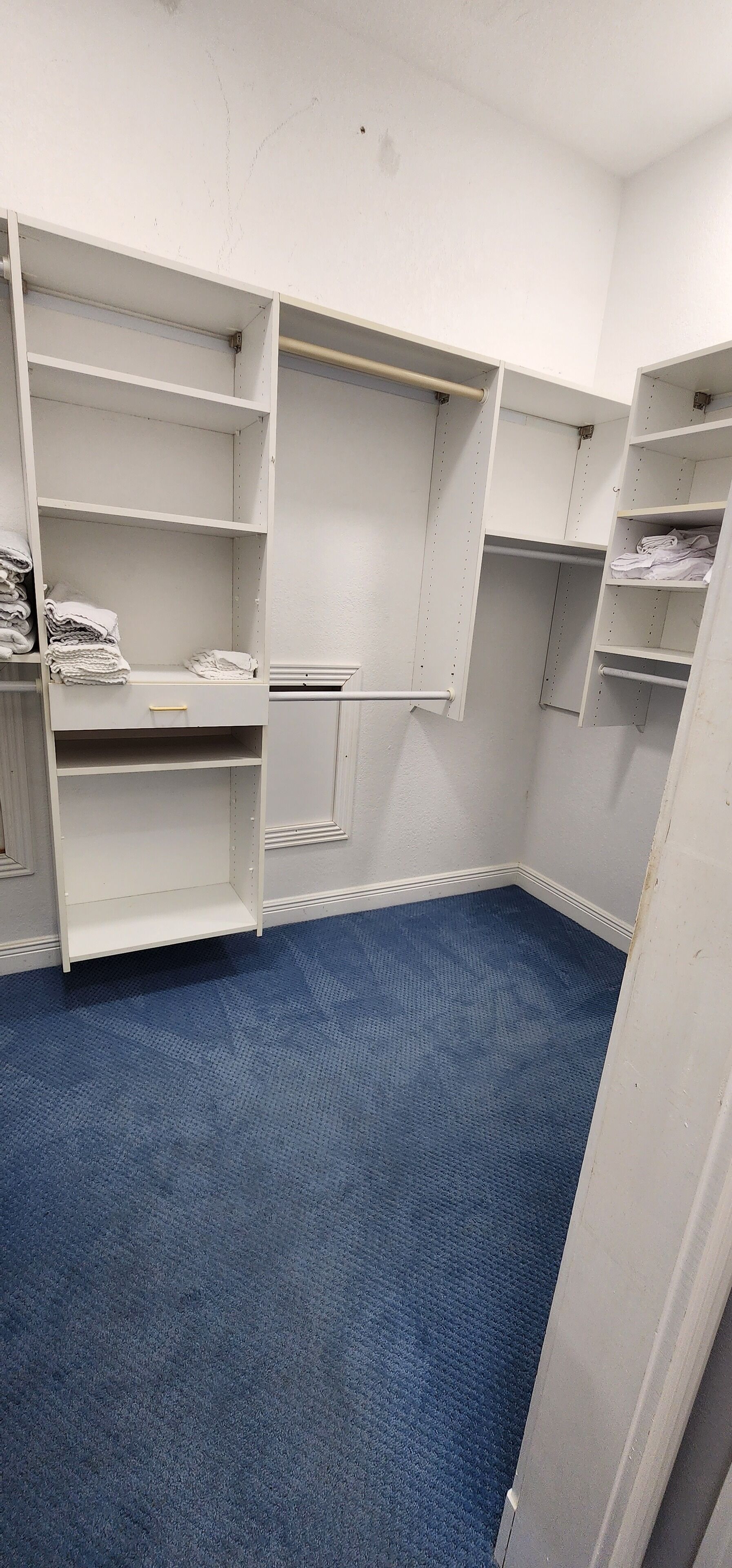 A room with a blue carpet, white walls, and a white wardrobe with multiple shelves and drawers.