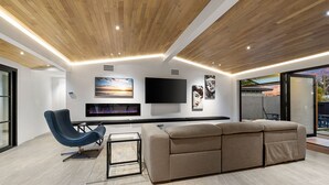 A modern living room with a wooden ceiling, a large flat-screen TV, a beige sofa, a blue chair, and artwork on the walls.