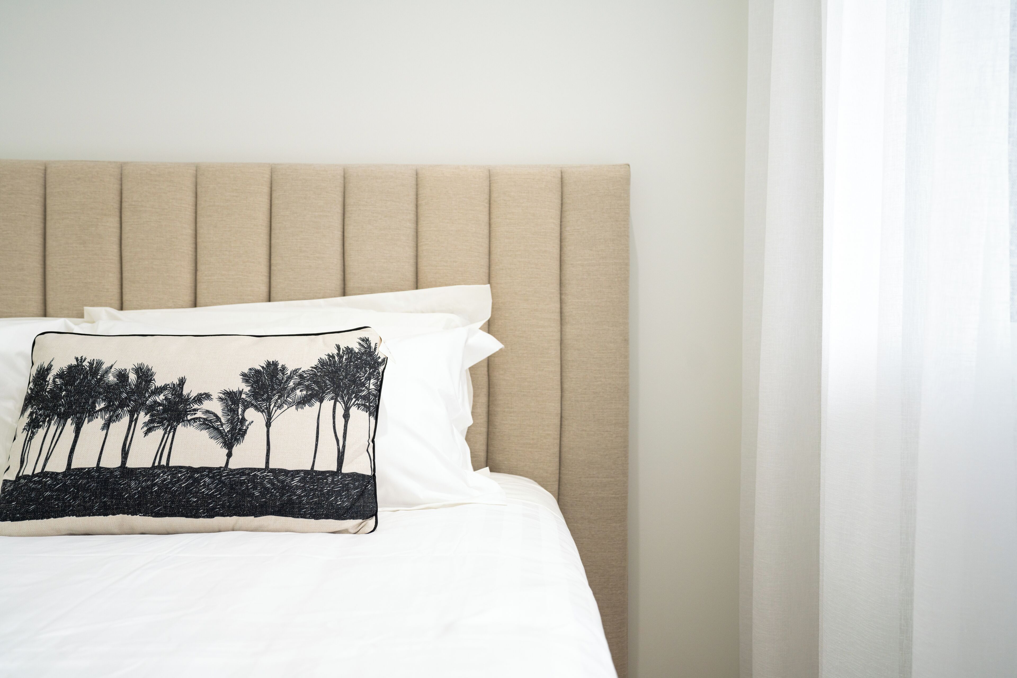 A bed with a beige upholstered headboard, white bedding, and a pillow with a palm tree design.