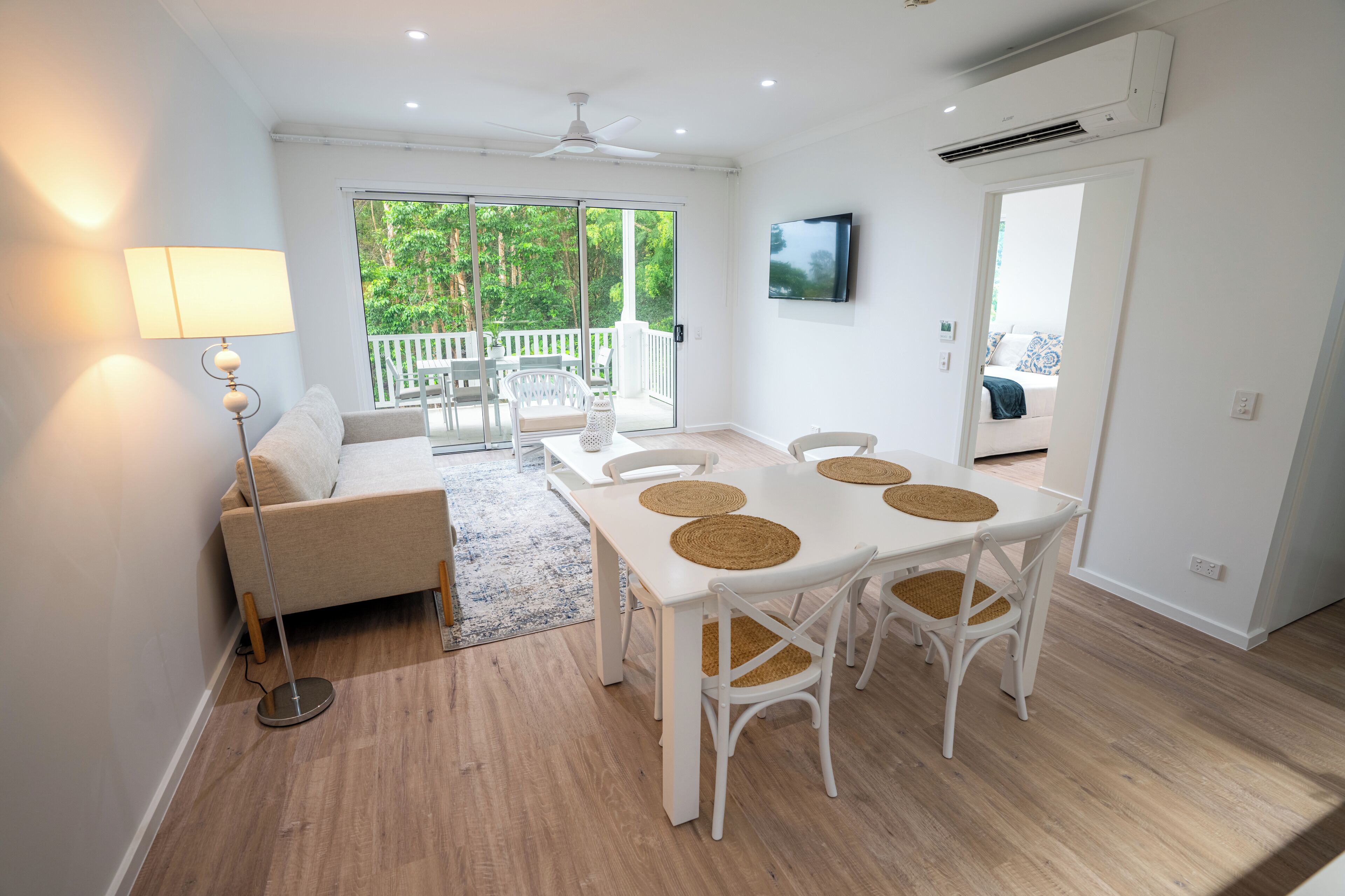 A modern, well-lit room with a dining table, chairs, a sofa, and a TV mounted on the wall.