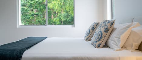 Un dormitorio con un ventanal grande, una cama con ropa de cama blanca y almohadas de estampado azul, y una manta oscura en el suelo.