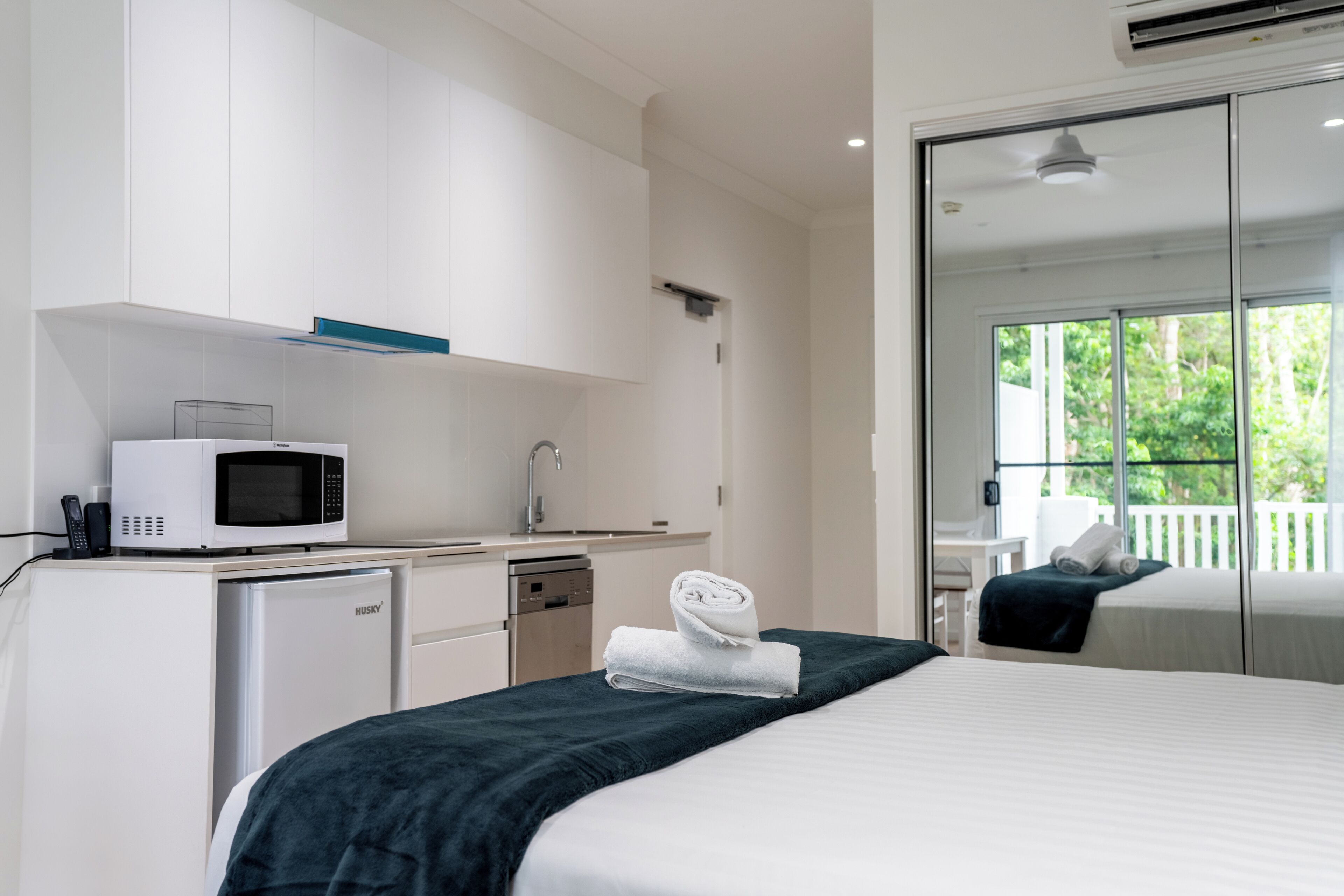A hotel room with a bed, a microwave, a small refrigerator, a sink, and a large window with a view of greenery.