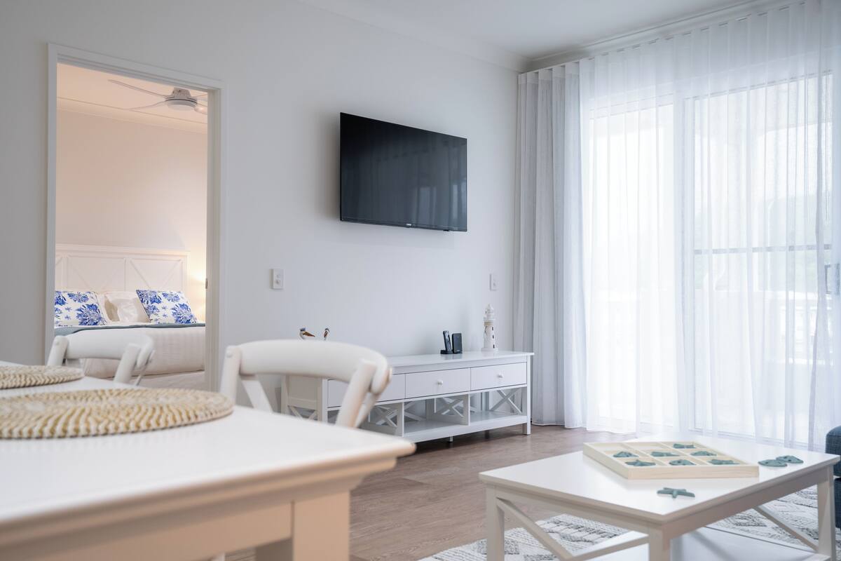 A modern, well-lit room with a flat-screen TV mounted on the wall, a white dining table with a placemat, a white sofa, and a white cabinet with drawers.